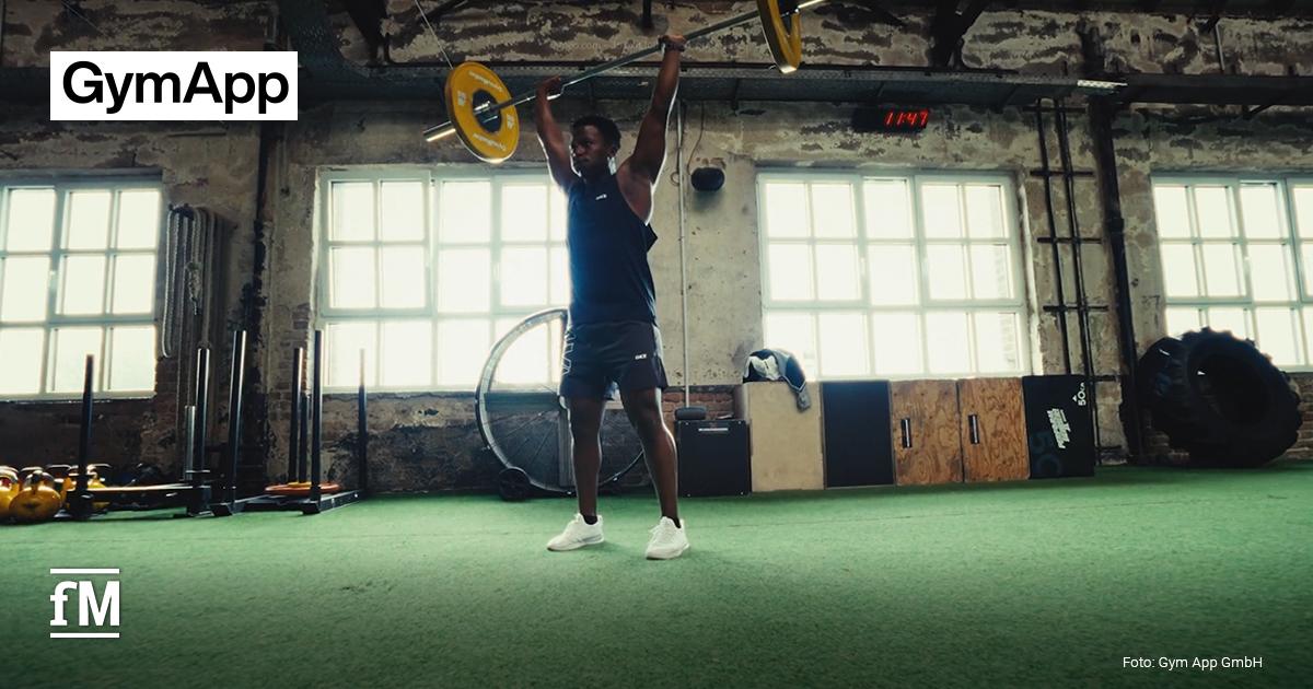 GymApp TranToGo im TV Innenraum eines Fitnessstudios mit grünem Kunstrasenboden; im Bildzentrum hebt ein Athlet eine Langhantel mit gelben Gewichtsscheiben über den Kopf; im Hintergrund große Fenster, Trainingsgeräte und Reifen; Kamera frontal auf Trainingsfläche