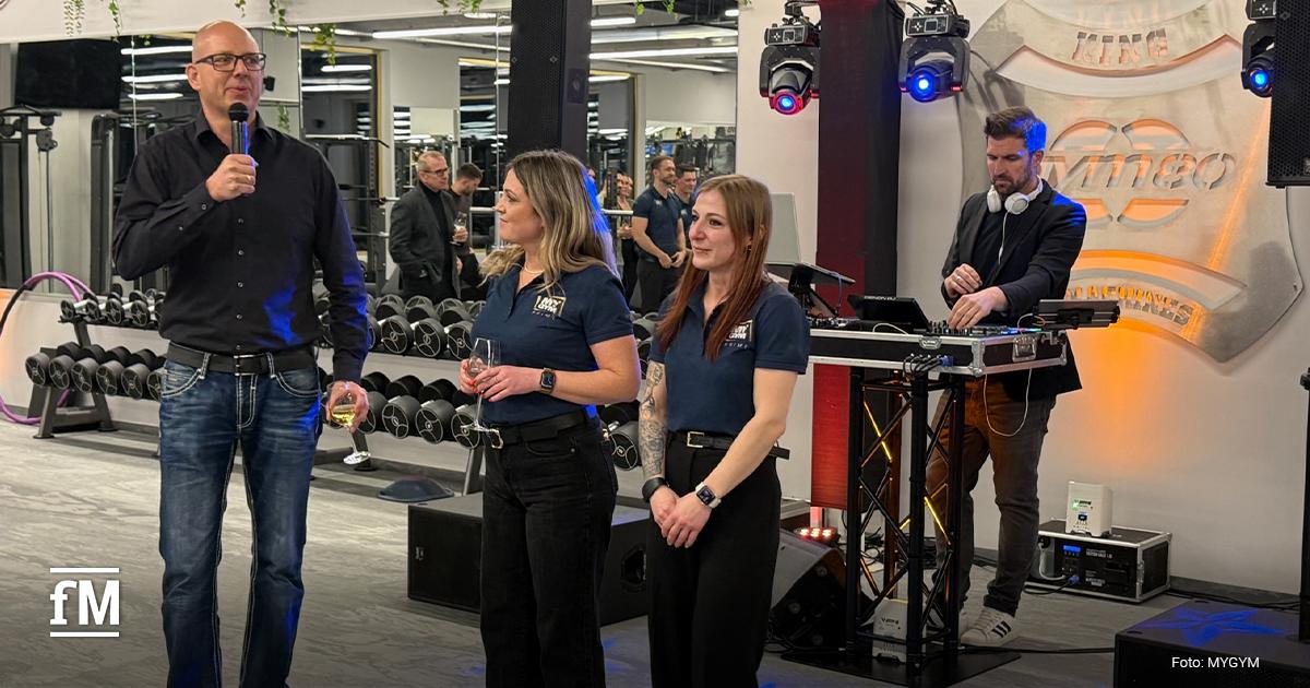 Innenraum eines Fitnessstudios mit Hantelregal im Hintergrund; vorne drei Personen, ein Mann mit Mikrofon spricht, zwei Frauen stehen daneben, rechts ein DJ an einem Mischpult mit Lautsprechern und Lichttechnik.