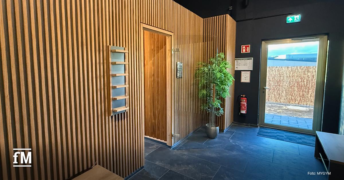 Innenraum im Eingangsbereich mit dunklem Fliesenboden, Holzlamellen an den Wänden, links eine Sitzbank, rechts eine Außentür mit Notausgangsschild und eine Pflanze im Topf