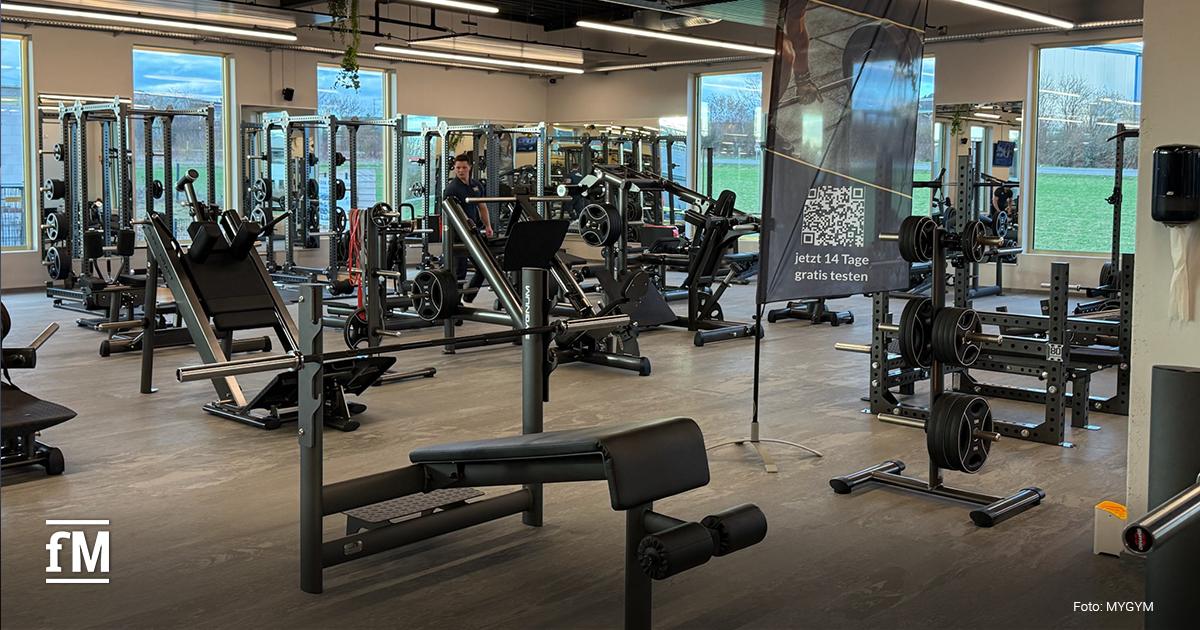 Trainingsfläche im Erdgeschoss mit Kraftgeräten, Langhantelstationen und Freihantelbereich; im Hintergrund Fensterfront mit Blick ins Grüne, rechts ein MYGYM PRIME Banner auf einem Aufsteller