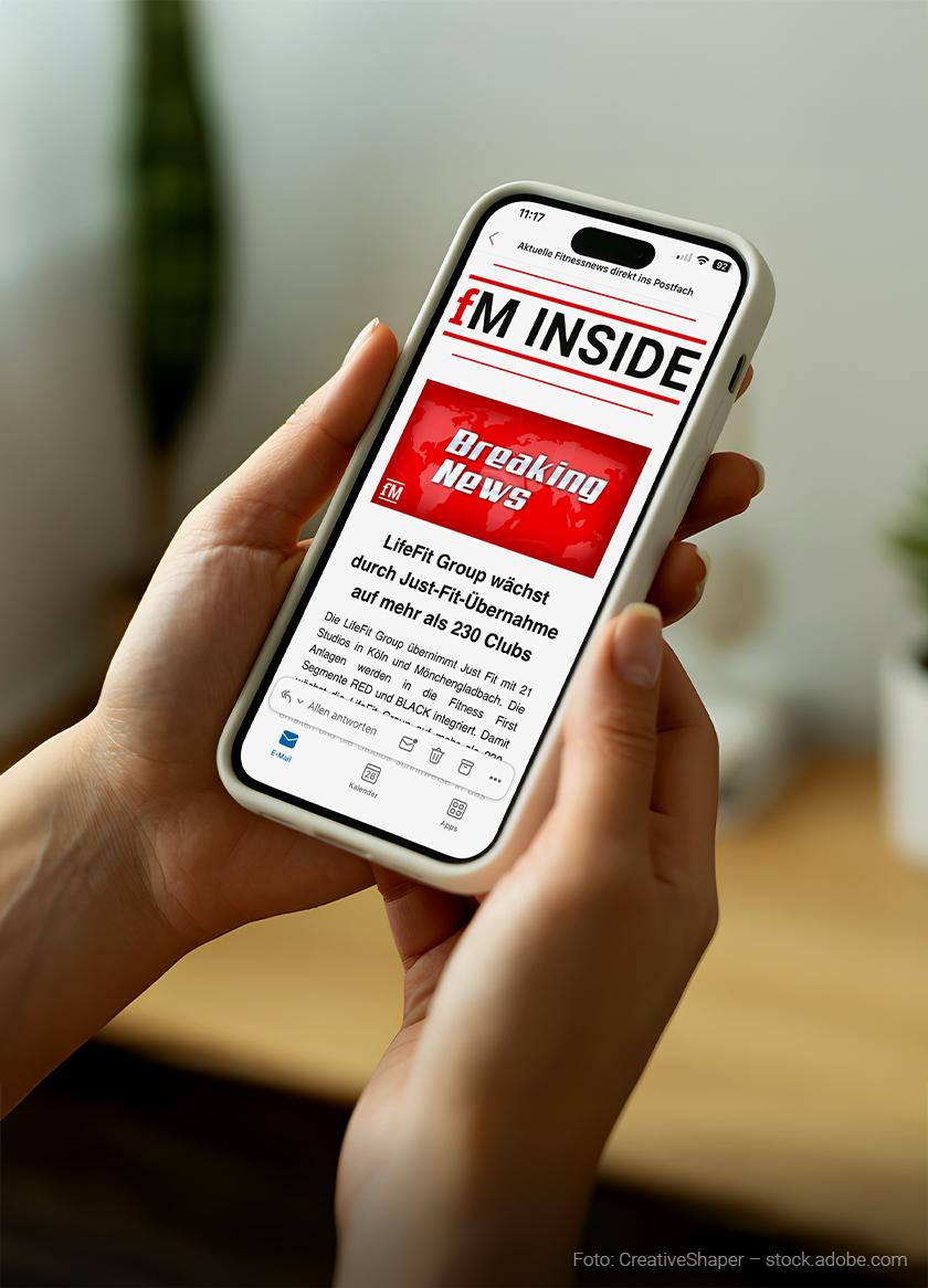 Zwei Hände halten ein Smartphone mit weißer Hülle. Auf dem Bildschirm ist ein geöffneter Newsletter mit dem Titel „fM INSIDE“ zu sehen. Darunter steht ein rotes Feld mit der Aufschrift „Breaking News“. Der Text darunter lautet „LifeFit Group wächst durch Just Fit-Übernahme auf mehr als 230 Clubs“. Im unteren Bereich sind typische E-Mail-Symbole wie Antworten, Weiterleiten und Löschen sichtbar. Im Hintergrund ist eine unscharfe Innenraumszene mit einer grünen Pflanze zu erkennen.