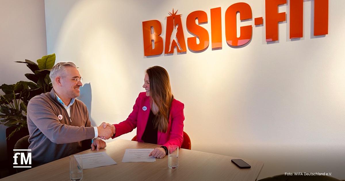 An einem hellen Holztisch sitzen sich zwei Personen gegenüber und geben sich die Hand. Auf dem Tisch liegen zwei Dokumente und zwei Wassergläser. Im Hintergrund befindet sich eine Wand mit großem orangefarbenem BASIC-FIT-Schriftzug, links steht eine Pflanze.