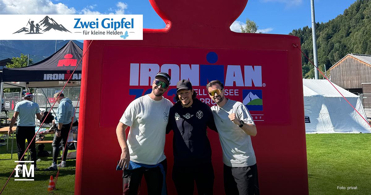 Zwei Gipfel für kleine Helden Drei Männer stehen vor einer großen roten Ironman-Figur auf einer Wiese, im Hintergrund Zelte, Berge und blauer Himmel