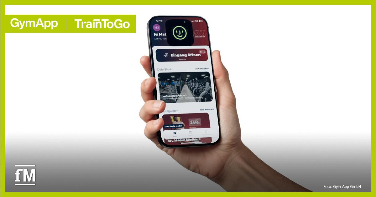 Studio-App TrainToGo Zentrale Vordergrundansicht eines Smartphones in einer Hand, Display zeigt die App-Oberfläche mit Button „Eingang öffnen“, Bereich „Dein Studio“ mit Bild eines Fitnessraums, Menüleiste unten, oben links Schriftzug „GymApp | TrainToGo“, unten rechts Text „Foto: Gym App GmbH“.