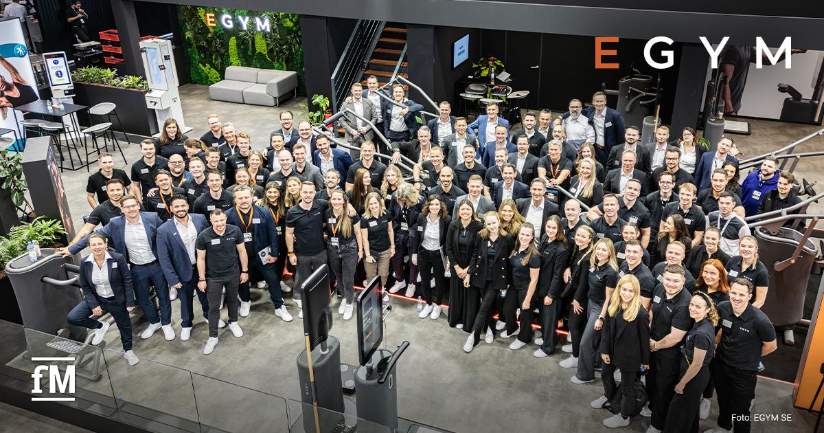 Große Gruppe von Mitarbeitenden in schwarzer oder dunkler Kleidung posiert auf einem Messestand mit Fitnessgeräten; oben steht groß „EGYM“.