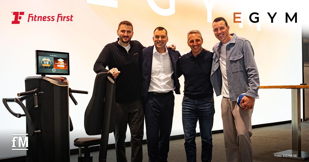 Im Vordergrund stehen vier Männer frontal neben einer schwarzen EGYM-Trainingsmaschine mit Sitz und Display: von links Daniel Kraus, Jan Scherzer, Martin Seibold und Dominik Klein. Links oben das rote Logo von Fitness First, rechts oben in der Ecke das von EGYM in Großbuchstaben, das E orangefarben, der Rest schwarz.