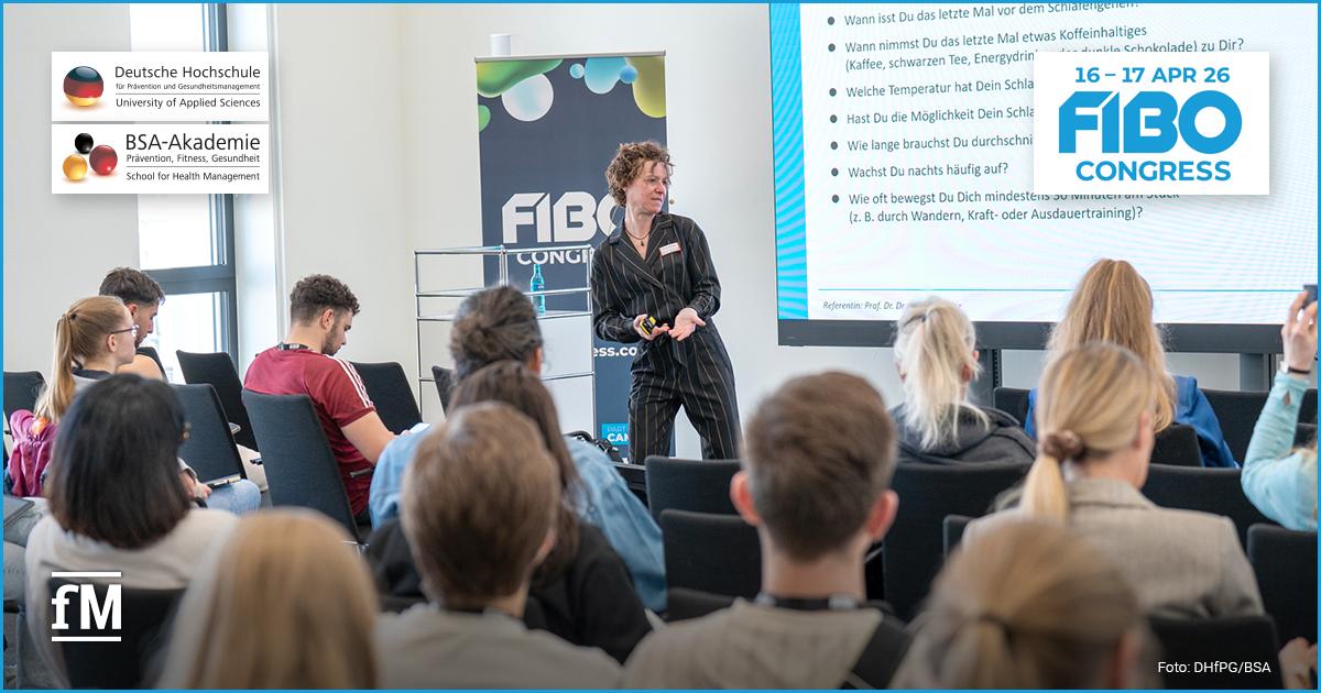 Vortragsszene in einem hellen Seminarraum: vorne eine Moderatorin mit kurzem lockigem Haar und schwarzem Anzug, die vor einem großen Bildschirm spricht; mehrere Sitzreihen mit Teilnehmern sind im Vordergrund zu sehen.