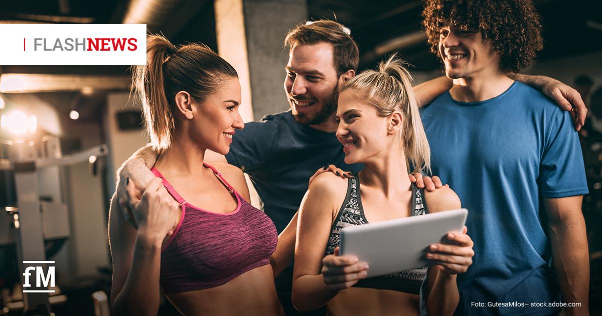 Vier junge Erwachsene in Sportkleidung stehen eng beieinander in einem Fitnessstudio, lächeln sich an und schauen auf ein Tablet; oben links steht „FLASHNEWS“.