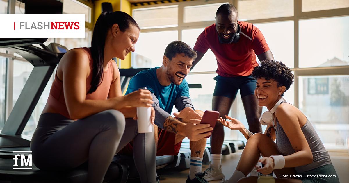 Vier lachende Fitnesssportler sitzen nach dem Training auf dem Boden eines Fitnessstudios und schauen gemeinsam auf ein Smartphone; neben ihnen stehen Trinkflaschen, im Hintergrund sind Laufbänder und große Fenster zu sehen.