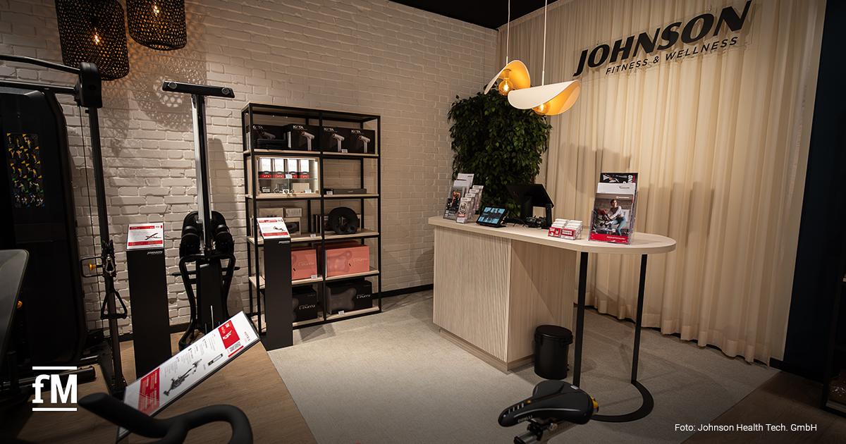 Empfangsbereich eines Johnson Fitness & Wellness Stores mit heller Theke rechts, Johnson Fitness & Wellness Logo auf einem Vorhang im Hintergrund, Produktregal mittig, Trainingsgerät und Infotafeln links im Vordergrund.