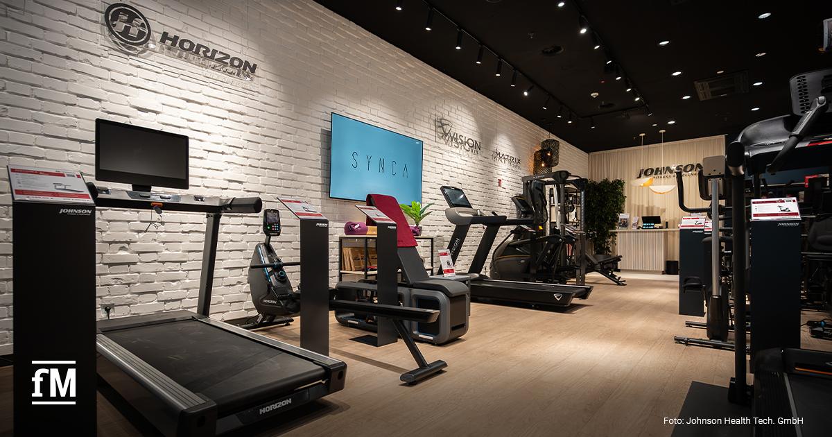 Innenraum eines Johnson Fitness & Wellness Stores mit Laufband links im Vordergrund, Produktdisplays und Infotafeln entlang einer weißen Backsteinwand, Bildschirm mit Synca Schriftzug mittig, Horizon Fitness Logo links oben, Vision Fitness und Matrix Fitness Logos mittig hinten, Johnson Fitness & Wellness Logo im hinteren Empfangsbereich.