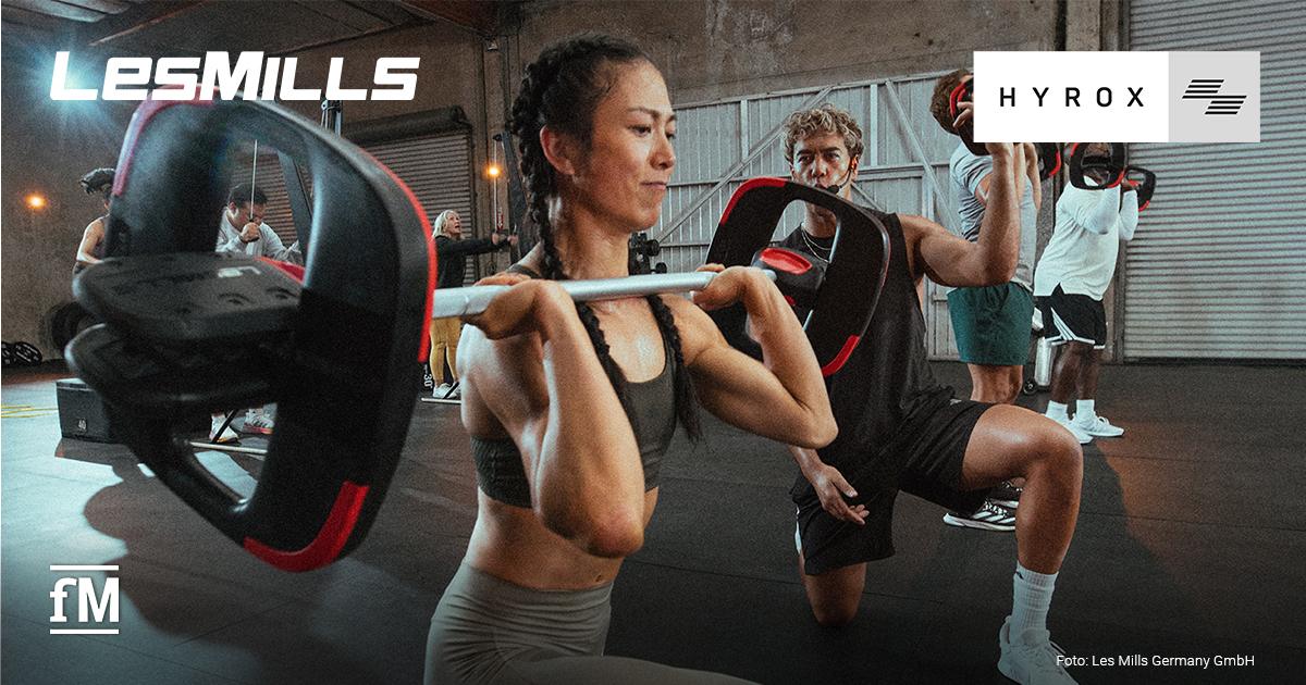 Frau im Vordergrund führt einen Ausfallschritt mit Langhantel aus, im Hintergrund mehrere trainierende Personen in einer Industriehalle.