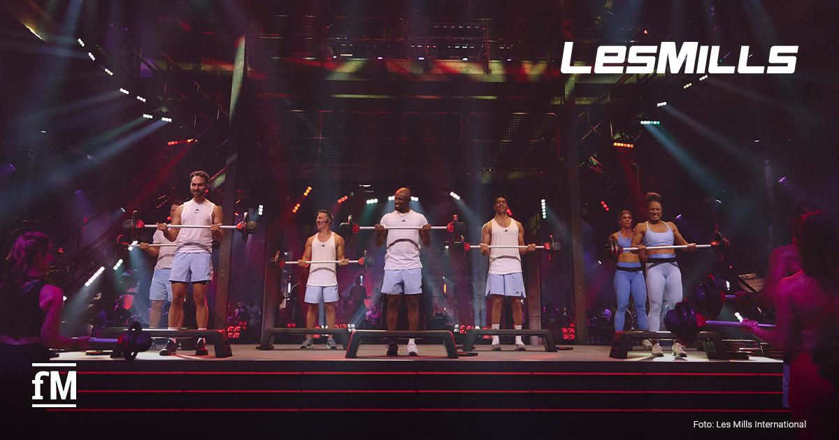 Mehrere Fitnesstrainer*innen stehen auf einer beleuchteten Bühne und trainieren mit Langhanteln vor Publikum. Oben rechts steht das Logo „LES MILLS“, die Szene wirkt wie eine große Liveshow mit Konzertlicht.