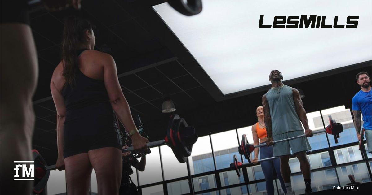 Les Mills Krafttraining Studie Mehrere Personen trainieren in einem Fitnessstudio mit Langhanteln. Links steht eine Person im Vordergrund mit dem Rücken zur Kamera, rechts trainieren drei Personen vor großen Fenstern. Oben rechts ist das Les Mills Logo zu sehen.