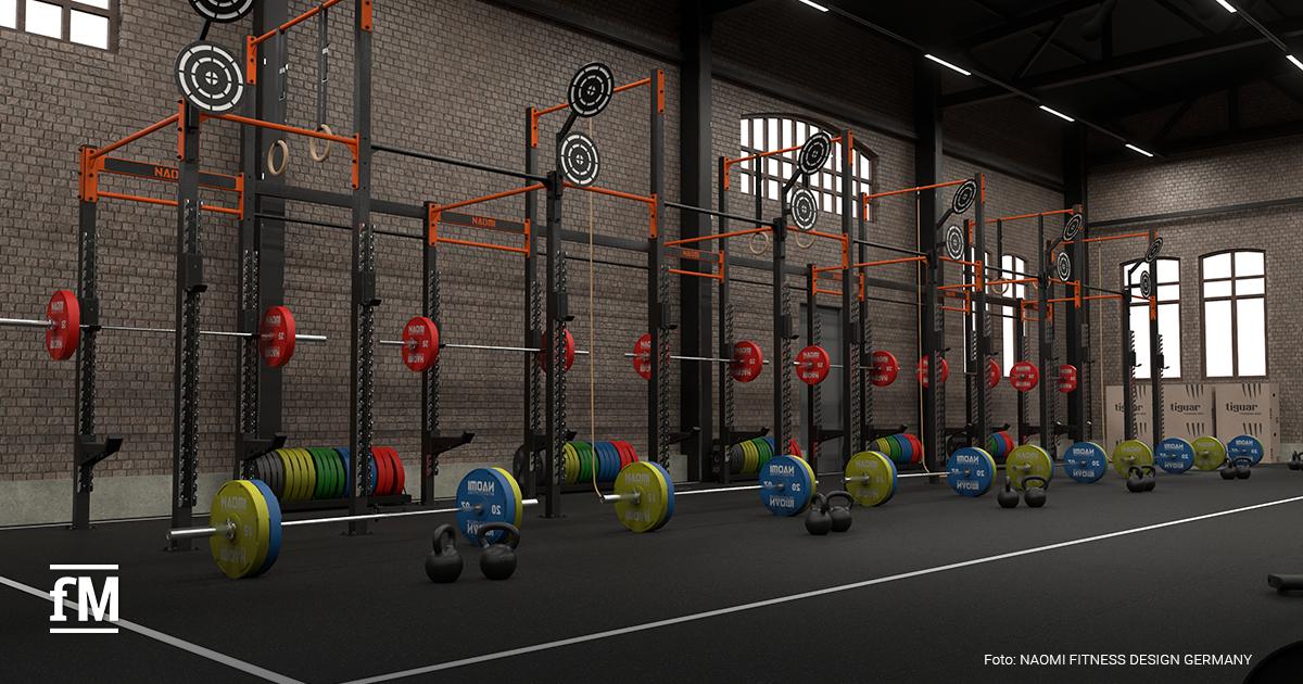 Reihe von Racks mit Langhanteln und farbigen Gewichtsscheiben im Vordergrund, Kettlebells am Boden, Zielscheiben an den Racks, Backsteinwand und Fenster im Hintergrund.