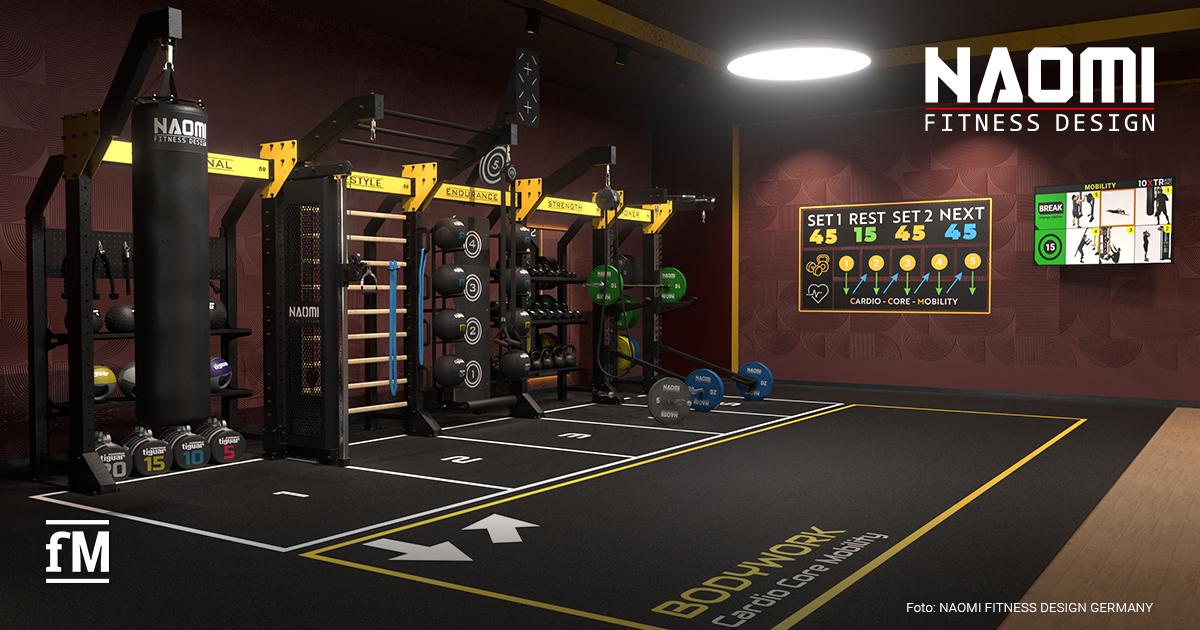 NAOMI FITNESS DESIGN FIBO 2026 Trainingsbereich mit Boxsack links vorne, Functional-Rig mit Hanteln und Medizinbällen mittig, Bodenmarkierungen mit Pfeilen und Schrift „BODYWORK Cardio Core Mobility“, Bildschirm mit Trainingsanzeige an der Wand.