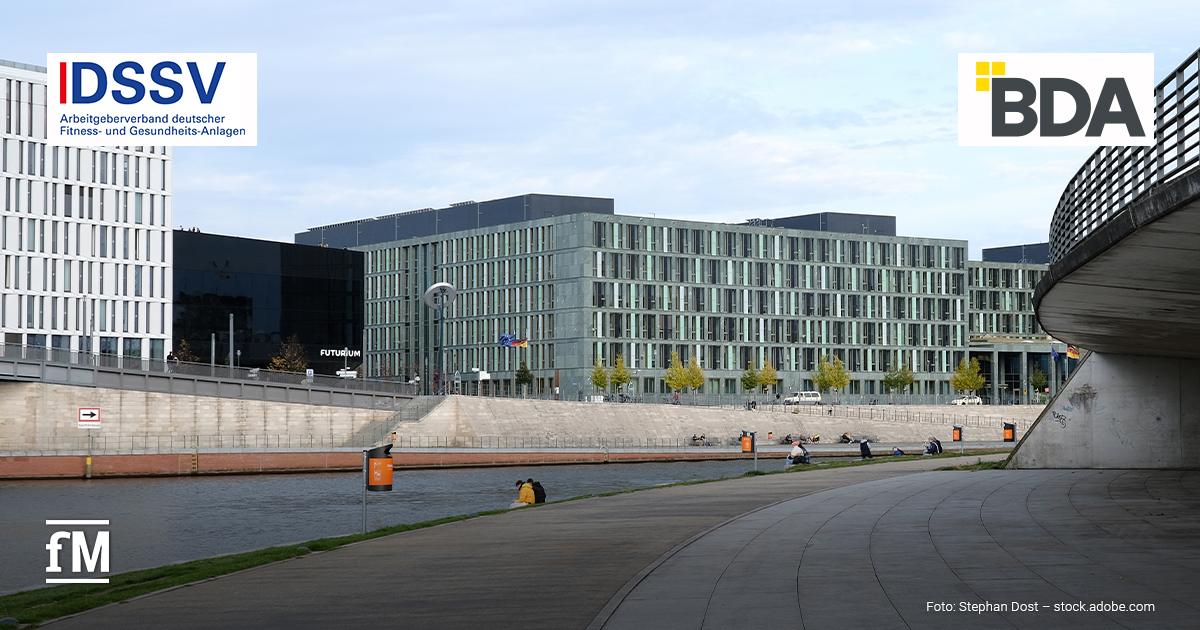 Uferpromenade an einem Fluss mit moderner Büroarchitektur im Hintergrund, links das Logo des „DSSV Arbeitgeberverband deutscher Fitness- und Gesundheits-Anlagen“, rechts das Logo der „BDA“, unten links der Schriftzug „fM“, unten rechts der Bildnachweis „Foto: Stephan Dost – stock.adobe.com“.