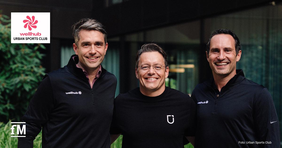 Drei lächelnde Männer stehen eng nebeneinander in einem begrünten Innenhof vor einer modernen Glasfassade. Alle tragen dunkle Oberteile, die beiden äußeren Männer tragen blaue Jeans.