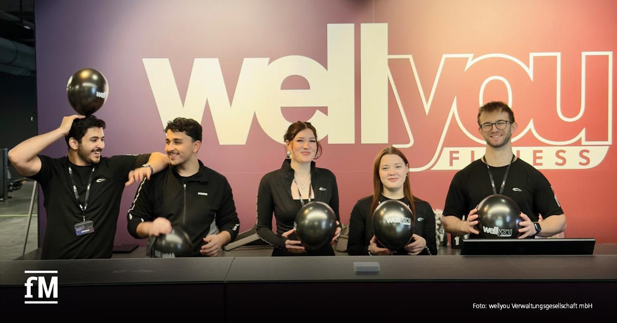 wellyou Mitarbeiter-App Fünf Mitarbeitende von wellyou stehen hinter einem Empfangstresen vor einer großen „wellyou Fitness“-Wand und halten schwarze Luftballons mit „wellyou“-Logo in den Händen. Links und rechts tragen die Personen schwarze Arbeitskleidung mit Namensbändern.