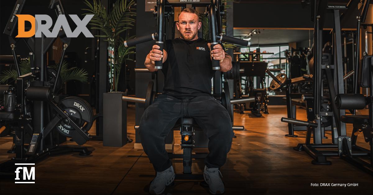 Im Vordergrund sitzt ein Mann an einem schwarzen Kraftgerät in einem Fitnessstudio und hält zwei Griffe auf Brusthöhe. Links oben steht das orange-graue Logo „DRAX“, links unten das weiße Logo „fM“, rechts unten der Text „Foto: DRAX Germany GmbH“.