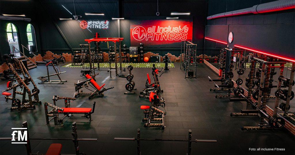 Großer Fitnessraum aus erhöhter Perspektive mit roten und schwarzen Kraftgeräten im Vordergrund, Hantelstangen, Trainingsbänken und Gewichten auf schwarzem Boden, grüner Laufbahn im hinteren Bereich, dunklen Wänden, roten Lichtlinien und leuchtenden Logos, beide mit der Aufschrift „all inclusive FITNESS“.