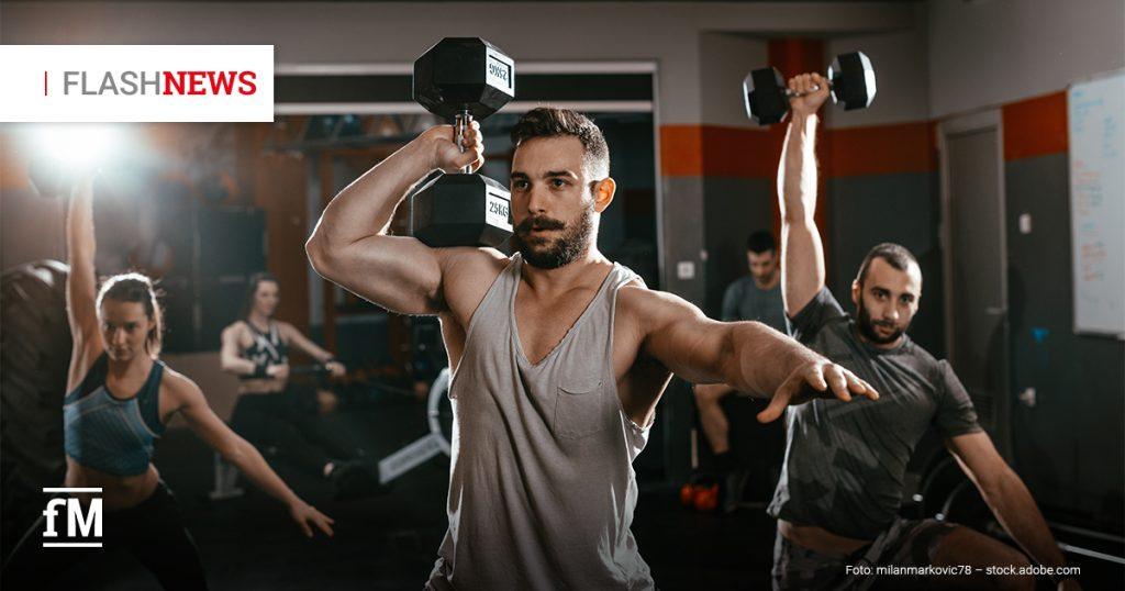 Muskelöser Mann im Fitnessstudio hebt eine Kurzhantel über den Kopf; im Hintergrund trainieren weitere Personen mit Gewichten.