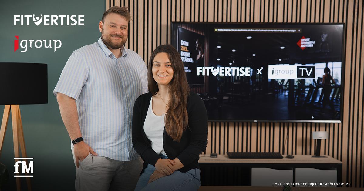 igroupTV und Fitvertise Kooperation Zwei Personen stehen vor einem Bildschirm mit den Logos „FITVERTISE“ und „igroupTV“. Im Vordergrund befindet sich ein Holztisch. Hinter den Personen verläuft eine Wand mit vertikalen Holzleisten und einem großen Monitor.