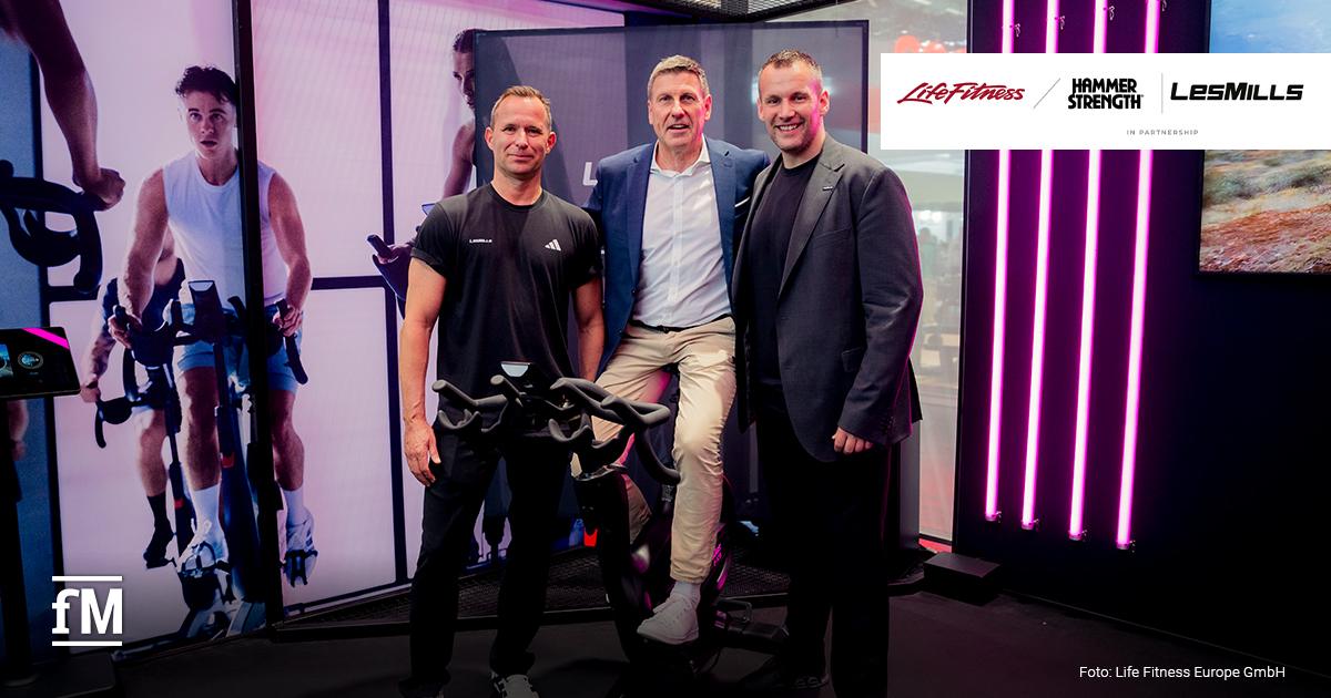 Life Fitness und Les Mills Im Vordergrund stehen zwei Männer neben einem Indoor Bike; ein dritter Mann sitzt auf dem Sattel. Der linke Mann trägt ein schwarzes Shirt mit den Schriftzügen „LES MILLS“ und „adidas“. Links im Hintergrund zeigt eine große Wandgrafik eine Person auf einem Bike, rechts stehen vier vertikale pinke Lichtleisten vor einer dunklen Wand.