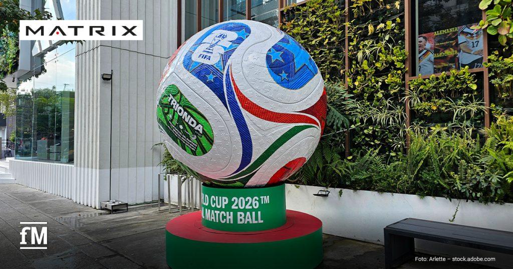 Vor einem Gebäude steht im Vordergrund ein großer Fußball auf einem grünroten Sockel mit den lesbaren Aufschriften „TRIONDA“, „FIFA“ und „WORLD CUP 2026™ OFFICIAL MATCH BALL“; rechts befinden sich Pflanzen, eine Sitzbank und ein Fenster mit Plakaten.