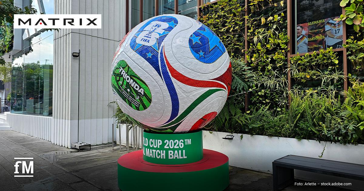 Matrix für den DFB: Volker Lichte Vor einem Gebäude steht im Vordergrund ein großer Fußball auf einem grünroten Sockel mit den lesbaren Aufschriften „TRIONDA“, „FIFA“ und „WORLD CUP 2026™ OFFICIAL MATCH BALL“; rechts befinden sich Pflanzen, eine Sitzbank und ein Fenster mit Plakaten.