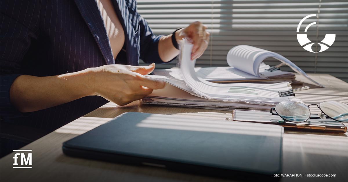 Sachverständige bei Bewertungsfragen in der Fitnessbranche Person blättert an einem Schreibtisch durch Unterlagen und Dokumente. Auf dem Tisch liegen Laptop, Brille und Aktenordner, während Sonnenlicht durch Jalousien fällt.