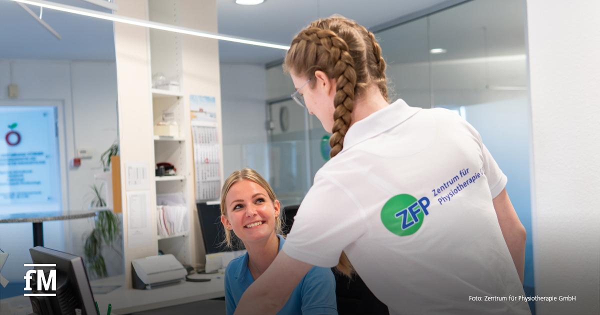 Zwei Frauen an einem Empfangstresen in hellen Praxisräumen. Eine Frau im weißen Polohemd mit ZFP-Logo und eine sitzende Frau im blauen Polohemd.