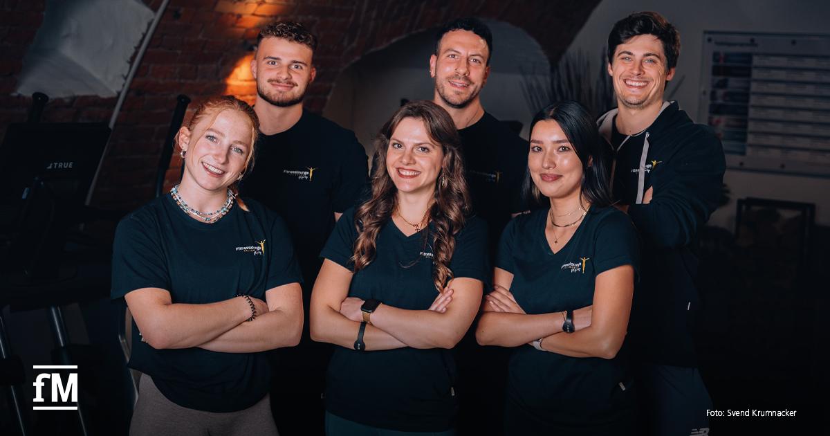 Sechs Mitarbeitende der Fitnesslounge Erlangen stehen in zwei Reihen vor einer Backsteinwand. Alle tragen dunkle Oberteile mit sichtbarem Fitnesslounge-Logo.