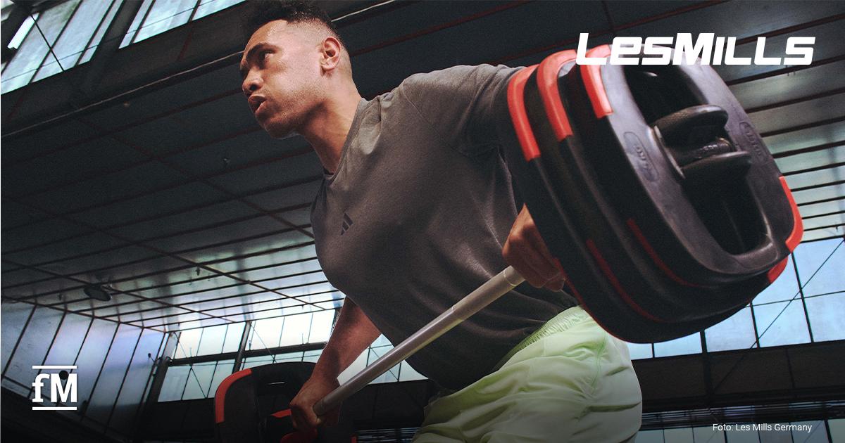 Les Mills Krafttraining Ein Mann trainiert im Vordergrund mit einer Langhantel mit schwarzen und roten Gewichtsscheiben in einem hellen Studio mit großen Fenstern; auf T-Shirt und Shorts stehen adidas-Logos.