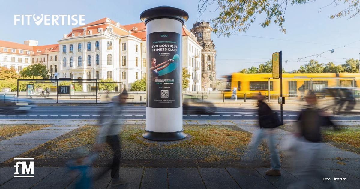 Advertorial Fitvertise Freistehende digitale Werbesäule im Vordergrund auf einem Platz am Straßenrand. Links oben im Bild steht das Logo „FITVERTISE“, links unten das Logo „fM“. Im Hintergrund befinden sich historische Gebäude, eine gelbe Straßenbahn und eine Haltestelle. Unscharfe Passantinnen und Passanten bewegen sich links und rechts vor der Werbesäule.