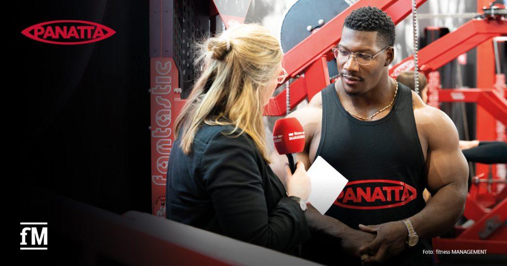 Eine Redakteurin von fitness MANAGEMENT interviewt den Bodybuilder Neckzilla an einem Panatta-Messestand. Im Hintergrund sind Trainingsgeräte zu sehen.