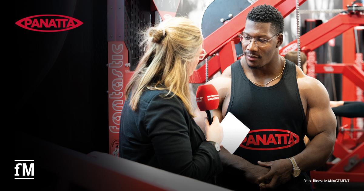 Interview Neckzilla Eine Redakteurin von fitness MANAGEMENT interviewt den Bodybuilder Neckzilla an einem Panatta-Messestand. Im Hintergrund sind Trainingsgeräte zu sehen.