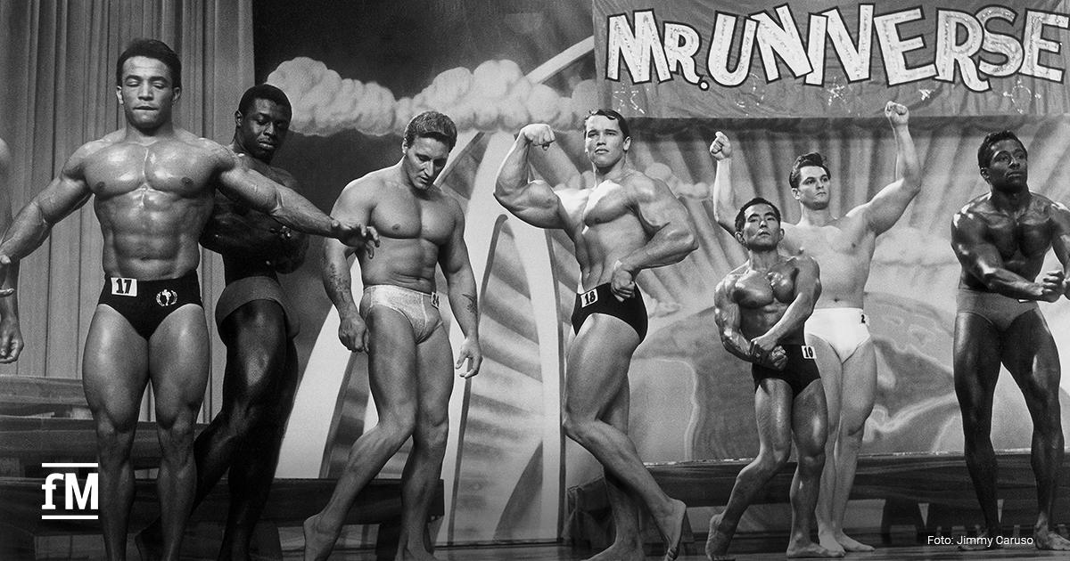 Mehrere Bodybuilder stehen auf einer Bühne und präsentieren Posen vor einem Bühnenbanner mit der Aufschrift „MR. UNIVERSE“.