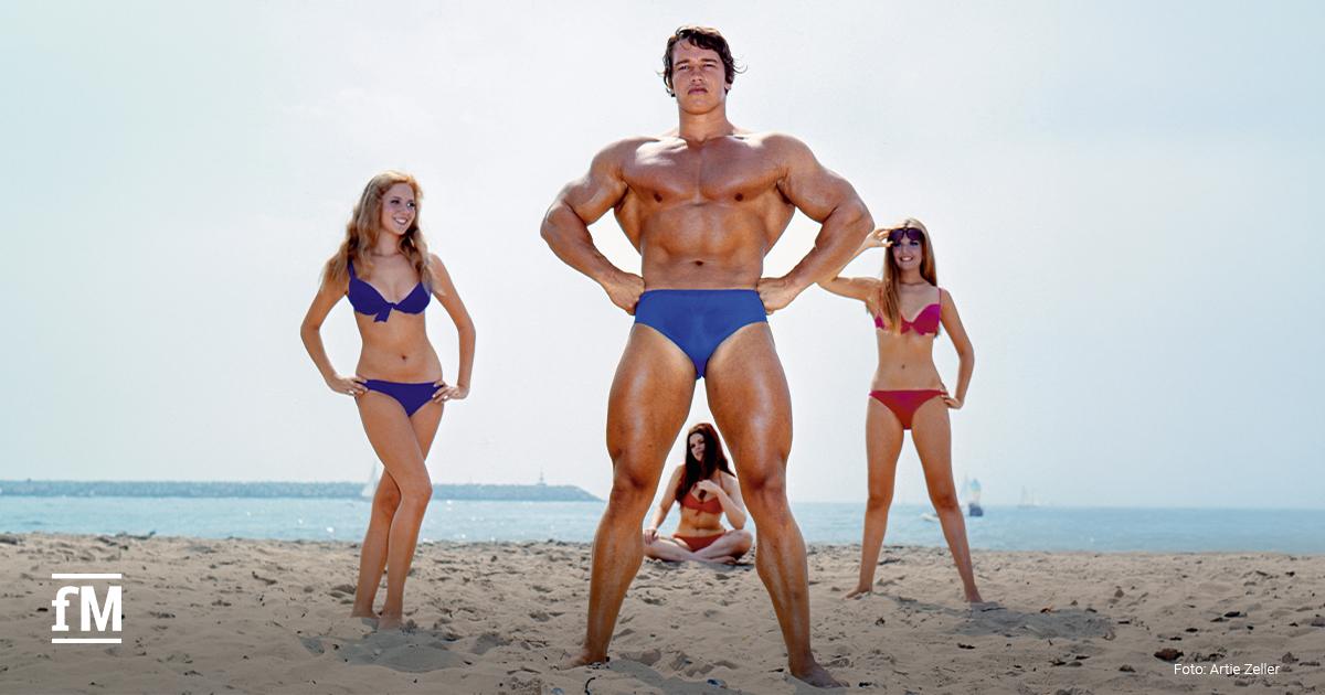 Muskelöser Mann in blauer Badehose steht am Strand, drei Frauen in Bikinis befinden sich im Hintergrund, Meer und Himmel dahinter.