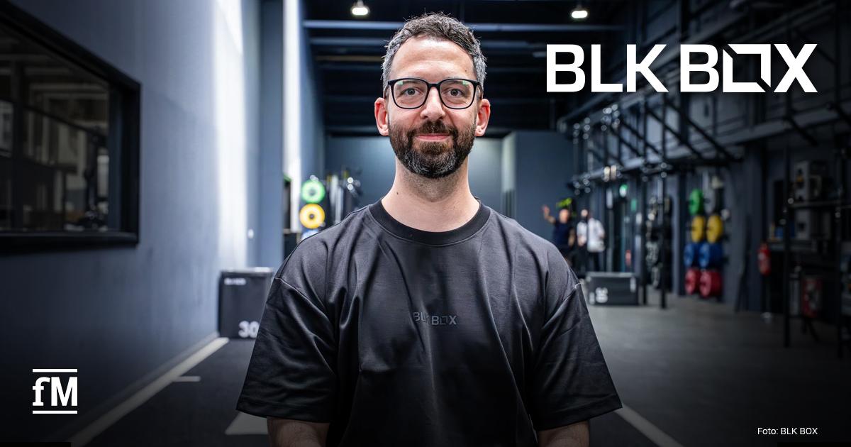 BLK BOX Timo Garrels Mann (BLK BOX Country Manager Timo Garrels) mit Brille und schwarzem T-Shirt steht in einer modernen Fitnesshalle vor Trainingsgeräten; rechts oben ist das BLK BOX-Logo zu sehen.