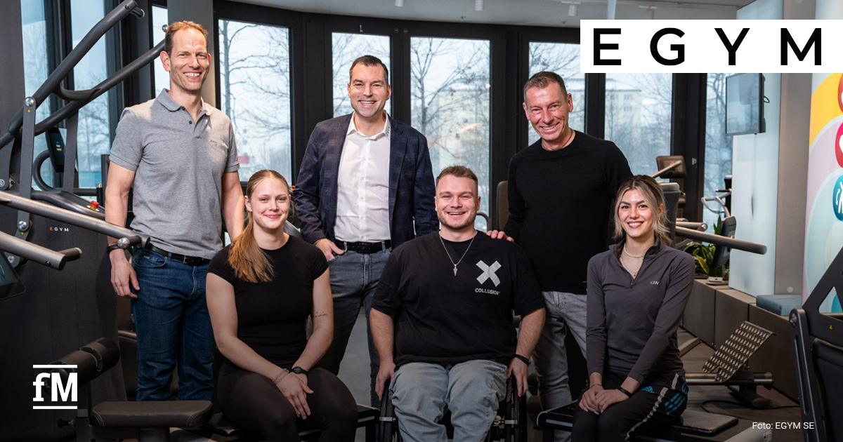 EGYM Future Champions Sechs Personen (v.l. Christian Tröger, Clara Hegemann, Jan Scherzer, Nicolai Sommer, Markus Othmer und Anya Atesli) posieren zwischen EGYM-Kraftgeräten für ein Gruppenfoto. Eine Person sitzt im Rollstuhl in der Bildmitte. Trainingsgeräte und Fenster im Hintergrund.