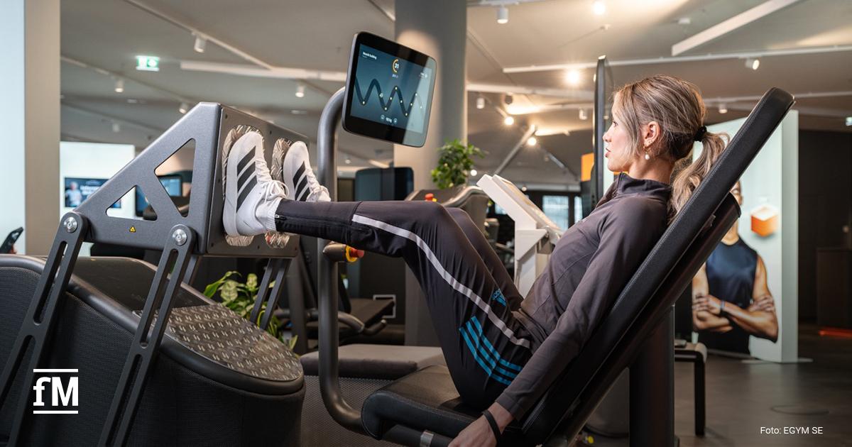 Frau sitzt an einem EGYM-Kraftgerät und blickt nach vorn. Im linken Vordergrund ein Monitor. Weitere Trainingsgeräte unscharf im Hintergrund