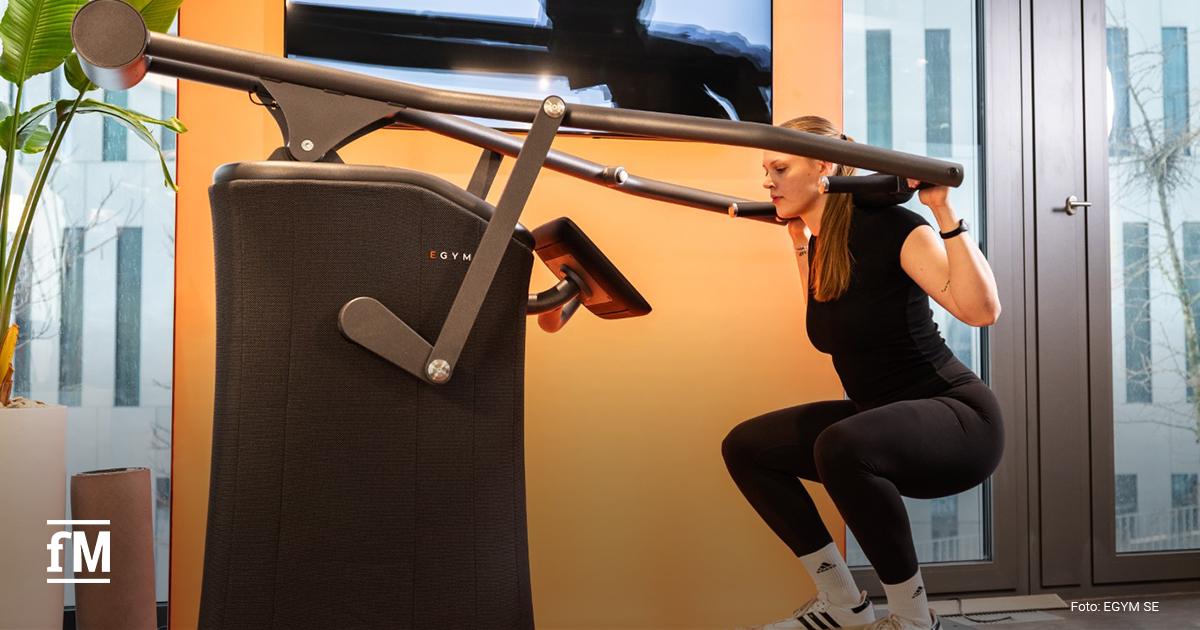 Frau führt eine Kniebeugenbewegung an einem EGYM-Gerät aus. Hände halten gepolsterte Griffe auf Schulterhöhe. Fenster und Bildschirm im Hintergrund.