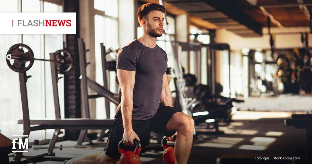 Im Vordergrund rechts trainiert ein Mann in schwarzer Sportkleidung in einem Fitnessstudio mit zwei Kettlebells in den Händen. Sein linkes Bein steht angewinkelt auf einer flachen Trainingsbank, sein rechtes Bein steht am Boden. Im Hintergrund links stehen Langhantelablagen vor hohen Fenstern, rechts liegen weitere Trainingsgeräte unscharf im Raum.