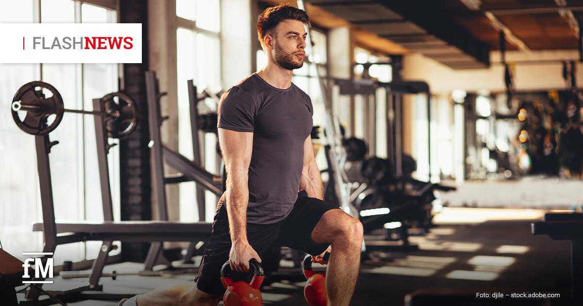 Fitness Flash News #06/2026 KW 25 Im Vordergrund rechts trainiert ein Mann in schwarzer Sportkleidung in einem Fitnessstudio mit zwei Kettlebells in den Händen. Sein linkes Bein steht angewinkelt auf einer flachen Trainingsbank, sein rechtes Bein steht am Boden. Im Hintergrund links stehen Langhantelablagen vor hohen Fenstern, rechts liegen weitere Trainingsgeräte unscharf im Raum.