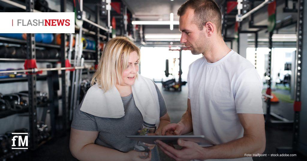 Eine Frau in Sportkleidung spricht mit einem Trainer, der ein Tablet hält. Beide blicken auf den Bildschirm. Im Hintergrund sind Krafttrainingsgeräte und Trainingszubehör zu sehen. Links oben ist das Logo „FLASHNEWS“ eingeblendet.