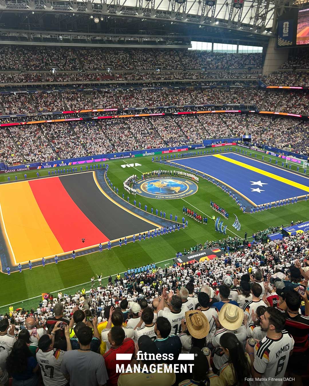 Aus hoher Stadionperspektive füllen Zuschauer die Ränge eines überdachten Fußballstadions. Auf dem Spielfeld liegen links eine große Deutschlandfahne und rechts eine große Curaçao-Flagge, dazwischen stehen Mannschaften und Offizielle um ein rundes FIFA WORLD CUP 2026 Emblem.