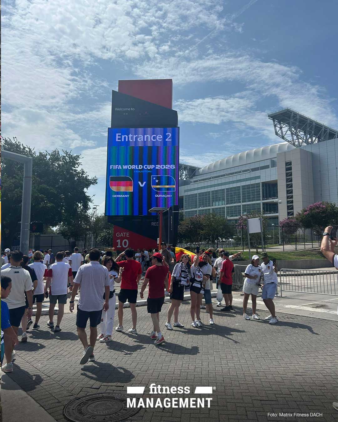 Vor einem Stadion stehen Menschen auf einem gepflasterten Vorplatz. Links der Mitte zeigt eine hohe digitale Tafel die Texte Entrance 2, FIFA WORLD CUP 2026, HOUSTON, GERMANY, CURAÇAO und Gate 10 mit den Flaggen Deutschlands und Curaçaos.