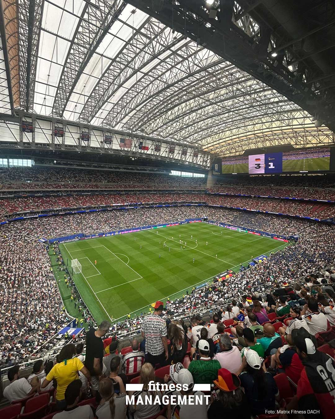 Aus erhöhter Tribünenperspektive blickt man in ein großes überdachtes Fußballstadion mit laufendem Spiel auf dem Rasen. Die Ränge sind dicht besetzt, oben rechts zeigt eine Stadionanzeige den Spielstand 3:1 mit Deutschlandflagge und Curaçao-Flagge.