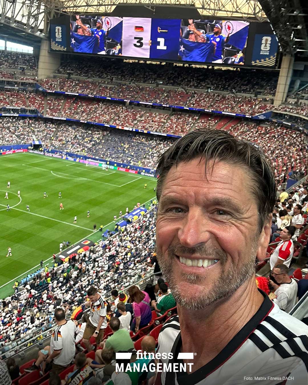 Rechts im Vordergrund nimmt eine lächelnde Person (Volker Lichte) ein Selfie auf einer Stadiontribüne auf. Links im Hintergrund läuft ein Fußballspiel auf dem Spielfeld, die Ränge sind dicht besetzt. Oben zeigt eine große Stadionanzeige den Spielstand 3:1 mit Deutschlandflagge und Curaçao-Flagge.